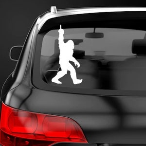 May include: White vinyl sticker of a Bigfoot silhouette with one arm raised in the air, applied to the rear window of a black car.
