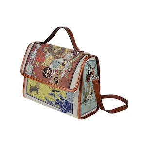 May include: A brown leather satchel handbag with a top flap closure and a long adjustable shoulder strap. The bag features a colorful illustration of characters from the Wizard of Oz, including Dorothy, the Scarecrow, the Tin Man, the Cowardly Lion, and Toto the dog.