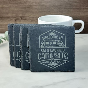 Puede incluir: Juego de cuatro posavasos de pizarra negra con un diseño blanco. El diseño presenta una autocaravana, árboles, una fogata y el texto "Bienvenido al camping de Saj & Laurie".