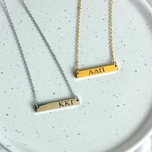 Könnte beinhalten: Zwei Halsketten mit Anhängern in Form von Stäben, eine silberne und eine goldene, jeweils mit eingravierten griechischen Buchstaben. Die silberne Halskette hat die Buchstaben KKT, und die goldene Halskette hat die Buchstaben ΑΔΠ. Beide Halsketten liegen auf einem weißen, gesprenkelten Teller.