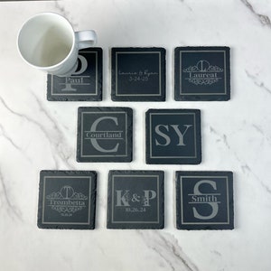 Op de afbeelding: Set van negen zwarte leisteen onderzetters met gegraveerde namen en data. De onderzetters zijn vierkant en hebben een licht getextureerd oppervlak.