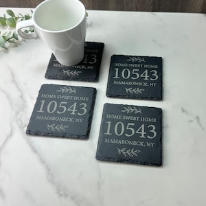 Op de afbeelding: Set van vier zwarte leisteen onderzetters met een wit gegraveerd ontwerp. Elke onderzetter heeft de tekst "Home Sweet Home" en het nummer "10543" met de stad en staat "MAMARONECK, NY".