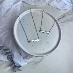 以下が含まれることがあります： 長方形のペンダントが付いた3つのシルバーネックレス。各ペンダントには「AXΩ」の文字が刻まれています。 ネックレスは白いセラミックプレートに展示されており、背景には白い布と緑があります。 チェーンは繊細で、ペンダントは小さいです。