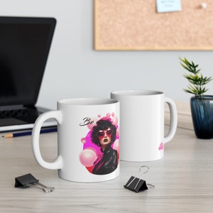 May include: White ceramic mug with a pink and black illustration of a woman wearing sunglasses and a leather jacket. The text "Be" is written in black cursive above the woman's head.