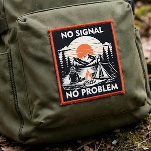 Puede incluir: Una mochila verde oliva con un parche rectangular de borde naranja. El parche muestra una escena de acampada en blanco y negro con una persona sentada junto a una hoguera, una tienda, montañas y un sol, con el texto NO SIGNAL NO PROBLEM.
