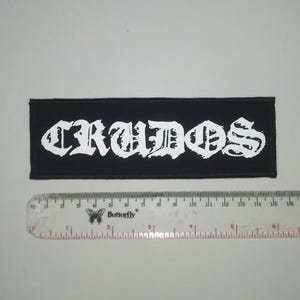 May include: Black and white embroidered patch with the word "CRUDOS" in a gothic font.