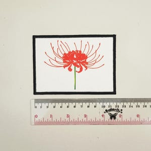 May include: A rectangular patch with a black border features a red spider lily illustration on a white background. The flower has thin, curved petals and a green stem. A clear ruler with centimeter markings is placed below the patch.