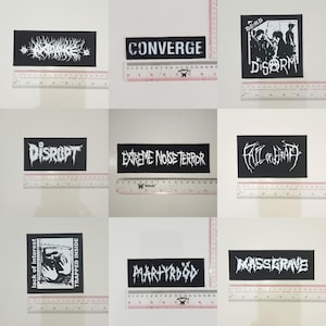 Peut inclure: Neuf écussons noirs avec texte blanc, affichant des noms de groupes tels que "Converge", "Disrupt" et "Extreme Noise Terror". Chaque écusson rectangulaire présente une typographie unique. Une règle est placée à côté de chaque écusson.