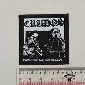 May include: Black and white patch with the text "CRUDOS" and "LAS INJUSTICIAS CAEN COMO PESADILLAS" in white lettering. The patch features two figures with their faces obscured.