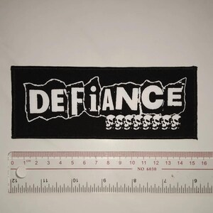 May include: Black and white embroidered patch with the word "DEFIANCE" in a bold, blocky font. A row of white skulls is below the word.
