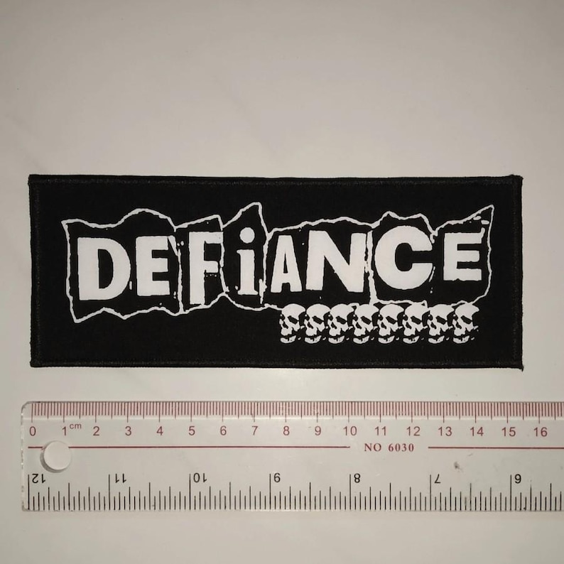 May include: Black and white embroidered patch with the word "DEFIANCE" in a bold, blocky font. A row of white skulls is below the word.