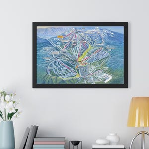 May include: Framed print of a ski resort map, showing colorful trails and lifts. The map is set against a backdrop of mountains and trees, with a black frame. The print is displayed on a white wall above a grey cabinet with decorative items.