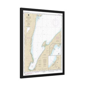 May include: A framed nautical chart of Keweenaw Bay, with detailed coastlines, depths, and navigational information. The chart features a beige background with blue and white details, encased in a black frame. The chart is a detailed map of the bay.