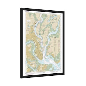 May include: A framed nautical chart of Charleston Harbor featuring detailed hydrographic data, depth soundings, and navigational markers in shades of blue, tan, and green, enclosed within a simple black rectangular frame.