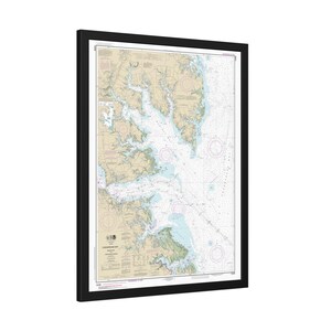 May include: A framed nautical chart of the Chesapeake Bay featuring detailed hydrographic data, depth soundings, and navigational markers. The map displays land areas in tan and water in light blue, enclosed within a simple black rectangular frame.