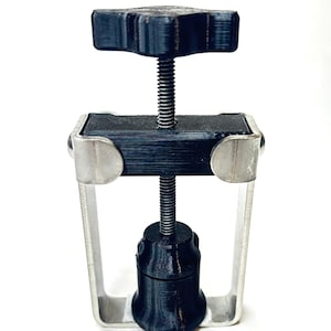 May include: A black and silver tool with a star-shaped knob at the top. The tool has a threaded rod and a base with a cylindrical component. The frame is silver, and the components are black.