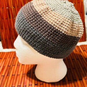 May include: A gray, brown, and beige crocheted beanie hat. The hat is on a mannequin head.
