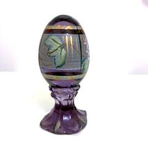 May include: A decorative purple glass egg on a matching stand. The egg features a floral design with gold accents and horizontal stripes. The stand has a flared base and a detailed, ornate design.
