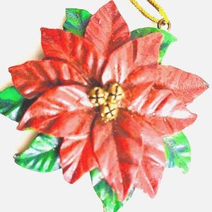 Op de afbeelding: Een kerstversiering van een poinsettia met rode bloemblaadjes en groene bladeren. Het midden van de bloem heeft drie kleine goudkleurige belletjes. Een gouden koord is bevestigd om op te hangen. Feestelijke vakantiedecoratie.