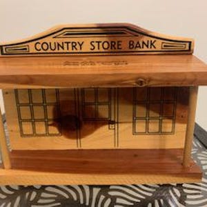 Può includere: Una "Country Store Bank" in legno con una facciata marrone chiaro e un tetto marrone scuro. La banca presenta un'illustrazione dettagliata di una facciata di negozio con finestre e una porta. Il testo "COUNTRY STORE BANK" è visualizzato in alto.