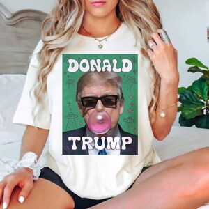 Op de afbeelding: Wit T-shirt met een groene afbeelding van Donald Trump die een bel blaast met de tekst "DONALD TRUMP" in een retro-lettertype.