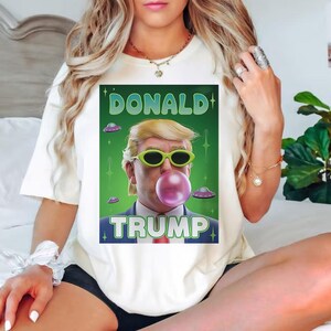Op de afbeelding: Wit T-shirt met een groene, witte en blauwe afbeelding van Donald Trump die een zonnebril draagt en een bel blaast. De afbeelding bevat ook de tekst "DONALD TRUMP" en verschillende vliegende schotels.