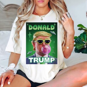 Op de afbeelding: Wit T-shirt met een groene, witte en zwarte afbeelding van Donald Trump met een zonnebril en een blaas. De tekst "DONALD TRUMP" staat op de afbeelding.