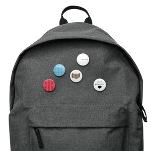 May include: A gray backpack with five different buttons pinned to it. The buttons are round and feature various designs, including a blue and white patterned button, a red button with white hearts, a button with a cartoon elephant, a white button with the text "Say Peanuts", and a black button with the text "You're phantastic" and a black heart.