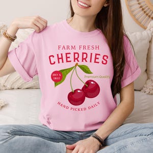 Könnte beinhalten: Hellrosa T-Shirt mit einer Grafik von zwei reifen Kirschen, grünen Blättern und dem Text "Farm Fresh Cherries". Zusätzlicher Text lautet "Juicy & Sweet", "Premium Quality" und "Hand Picked Daily".