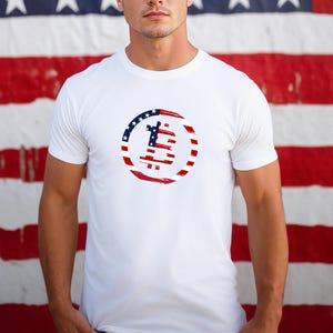 May include: White t-shirt featuring a Bitcoin symbol designed with red, white, and blue brushstrokes, resembling the American flag. The Bitcoin symbol is centered on the chest. The shirt has short sleeves and a crew neck.