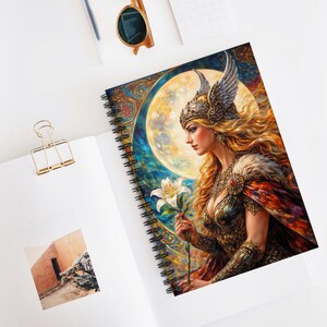 May include: Spiral-bound notebook featuring a detailed illustration of a woman with long blonde hair, wearing a winged helmet and holding a white lily. The artwork is set against a backdrop of a full moon and ornate, colorful patterns.