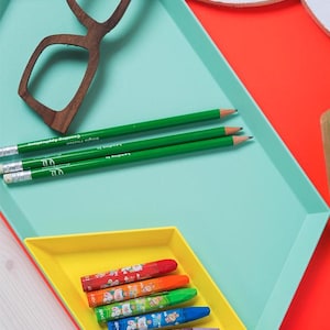 May include: A colorful still life featuring a wooden-framed pair of glasses, green pencils, and crayons. The items are arranged on a geometric tray with a teal, yellow, and red color scheme. The crayons are in a small yellow tray.