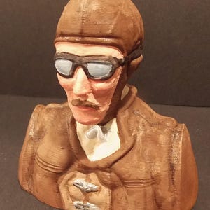May include: A brown bust of a person wearing a brown leather helmet and goggles. The person has a mustache and is wearing a brown leather jacket with silver buttons.