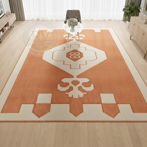 Könnte beinhalten: Ein orangefarbener und cremefarbener Teppich mit geometrischem Design. Der Teppich hat eine zentrale Diamantenform mit einem sich wiederholenden Muster, umgeben von einem cremefarbenen Rand. Der Teppich liegt auf einem hellen Holzboden.