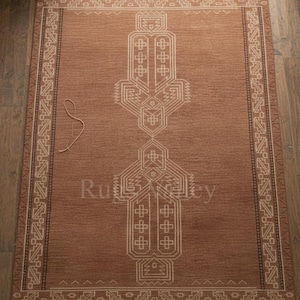 Könnte beinhalten: Ein rechteckiger brauner Teppich mit einem zentralen geometrischen Design in Beige und einem gemusterten Rand. Der Teppich trägt den Text "Rug Valley" in der Mitte. Der Teppich liegt auf einem Holzboden.