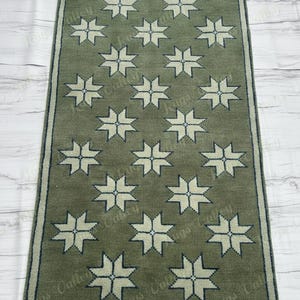 Könnte beinhalten: Ein rechteckiger, olivgrüner Teppich mit einem sich wiederholenden Muster aus cremefarbenen Sternenmustern. Der Teppich hat einen dunkelgrünen Rand und Fransen an den kurzen Enden. Er ist etwa 122 cm lang und 61 cm breit.