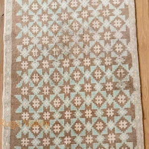 Könnte beinhalten: Ein rechteckiger Teppich mit geometrischem Muster. Der Teppich hat ein sich wiederholendes Design aus braunen, hellblauen und weißen Formen. Der Rand ist in einem helleren Braunton gehalten. Der Teppich liegt auf einem Holzboden.