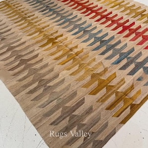Könnte beinhalten: Ein beiger Teppich mit einem geometrischen Muster in Rot-, Blau-, Gelb- und Brauntönen. Der Teppich hat ein sich wiederholendes Design aus abgewinkelten Formen, wodurch ein strukturiertes Aussehen entsteht. Der Text "Rugs Valley" ist in der unteren Ecke sichtbar.