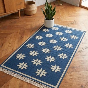 May include: A rectangular blue rug with a repeating white starburst pattern. The rug has a white border and fringe along the short ends. The rug is placed on a wooden floor.