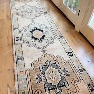 Könnte beinhalten: Ein langer Läufer mit cremefarbenem Grund und sich wiederholenden geometrischen Medaillonmustern in Schwarz, Grau, Beige und gedämpftem Orange. Die Bordüre zeigt eine Reihe kleiner quadratischer Motive in abwechselnden Farben an den Rändern.