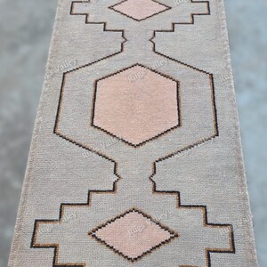 Könnte beinhalten: Ein rechteckiger, hellgrauer Teppich mit geometrischem Muster. Das Design zeigt einen zentralen Sechseck und rautenförmige Formen in einem zarten Rosa, umrandet in Schwarz und Braun. Der Teppich hat Fransen an den kurzen Enden.