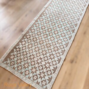 May include: A rectangular, patterned rug with a brown and light blue geometric design. The rug has a light beige border and is placed on a wooden floor. The pattern features star-like shapes and is suitable for a hallway or entryway.