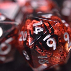 May include: Close-up of polyhedral dice with a red and black speckled design. The dice have silver numbers and a black spider graphic. The dice are used for tabletop games.