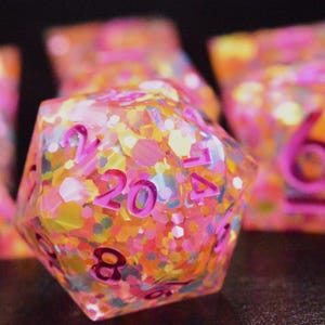May include: Close-up of a set of colorful polyhedral dice. The dice are filled with pink, yellow, and orange glitter. The numbers are in a bright pink color. The largest die shows the number 20.