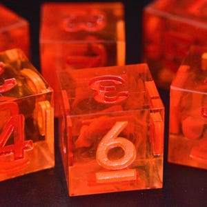 Op de afbeelding: Verschillende doorschijnende oranje dobbelstenen met rode cijfers zijn gerangschikt op een donkere ondergrond. De dobbelstenen zijn kubusvormig en de cijfers 1 tot en met 6 zijn zichtbaar op de vlakken. De dobbelstenen lijken van helder, glanzend materiaal te zijn.