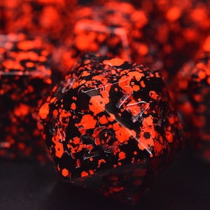 May include: Close-up of several black dice with vibrant red glitter embedded throughout. The dice have a glossy finish and are set against a dark background, highlighting the contrast between the colors. The dice are likely used for tabletop games.