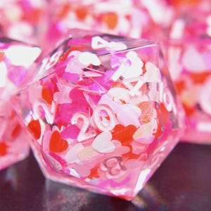 May include: Close-up of several clear, polyhedral dice filled with pink, red, and white heart-shaped confetti. The dice have white numbers and are set against a blurred background, creating a romantic theme.