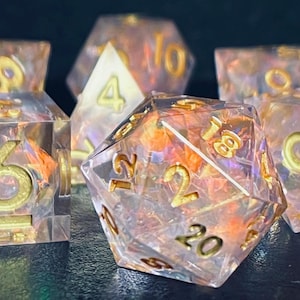 Op de afbeelding: Een set iriserende dobbelstenen met gouden cijfers. De dobbelstenen zijn helder en hebben een draaiende structuur van binnen. De cijfers op de dobbelstenen zijn 4, 6, 8, 10, 12, 18 en 20.