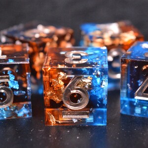 May include: A set of five translucent resin dice featuring a dual-tone blue and orange color scheme with embedded metallic gold flakes. Each cube-shaped die displays large, clear, recessed numerals on its faces, showcasing a sharp-edged design.