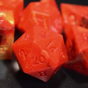 May include: A set of polyhedral dice in a vibrant red resin with swirling orange and black marbled patterns. The dice feature engraved numbers, including a twenty-sided die in the foreground showing the number twenty clearly visible on its face.